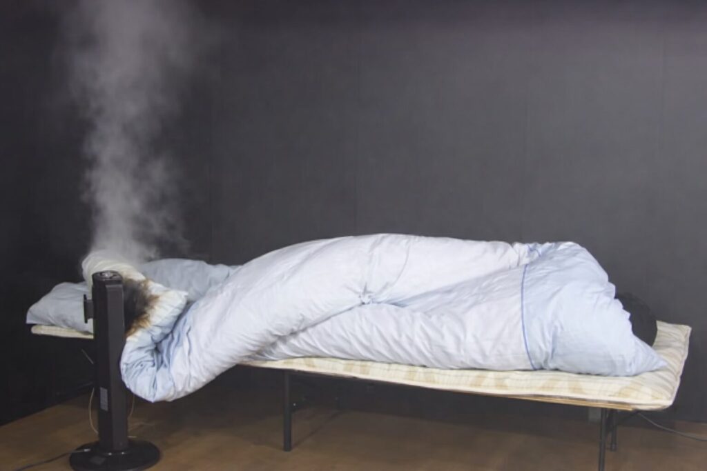 電気ストーブの吹き出し口が布団や枕に近接し、横向きで寝ている人の足元付近に温風や熱が直接当たっている、火災が起きる直前の危険な室内状況を写した写真。