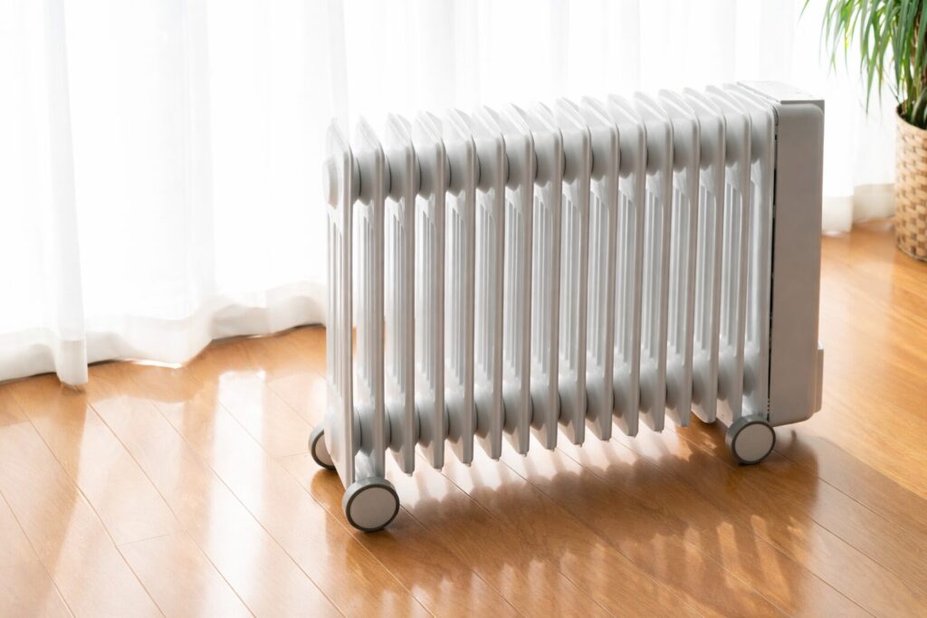 窓際のフローリング床にキャスター付きのオイルヒーター型電気ストーブが設置され、白いカーテン越しの自然光が当たっている室内の様子。