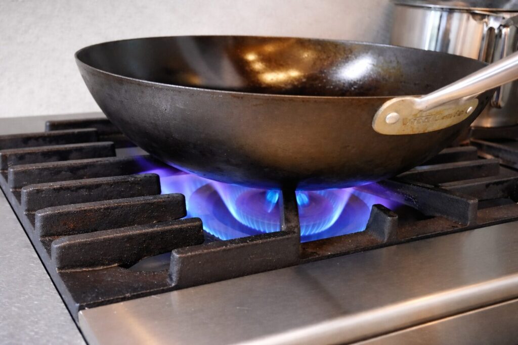 業務用ガスコンロの五徳の上で、中華鍋を強火で加熱している様子。鍋底の下には青いガスの炎が均一に広がり、使い込まれた鍋肌とステンレス製コンロの質感が確認できる構図。