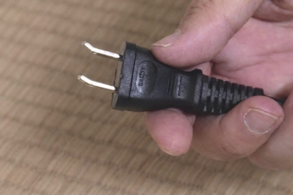 コンセントプラグの金属刃がわずかに曲がった状態で、手に持って先端部分を確認している様子。黒色の電源プラグと木目調の床面が写っている。