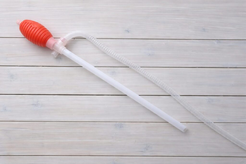 赤いゴム製のポンプ部分と白いパイプ、透明なホースで構成された灯油用サイフォンポンプが、明るい木目の床の上に横向きに置かれている様子。