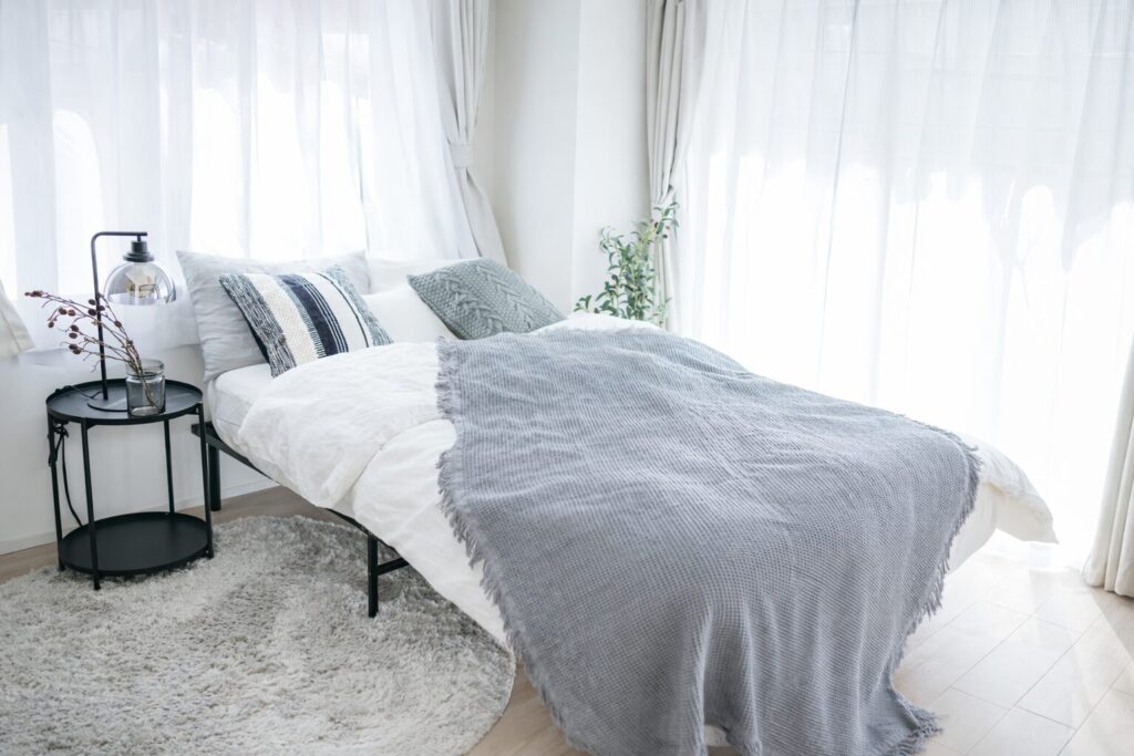 白いカーテン越しに自然光が差し込む明るい寝室で、白いベッドリネンとグレーのブランケットが整えられ、横に黒いサイドテーブルと観葉植物が配置されている様子。