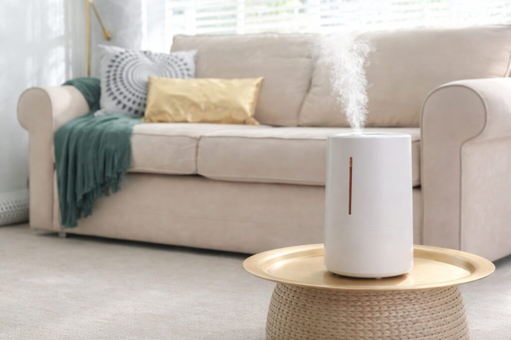 白を基調としたリビングルームで、丸いサイドテーブルの上に置かれた円筒形の加湿器から白い蒸気が立ち上り、背後にはクッションとブランケットのあるソファが配置されている落ち着いた室内の様子。