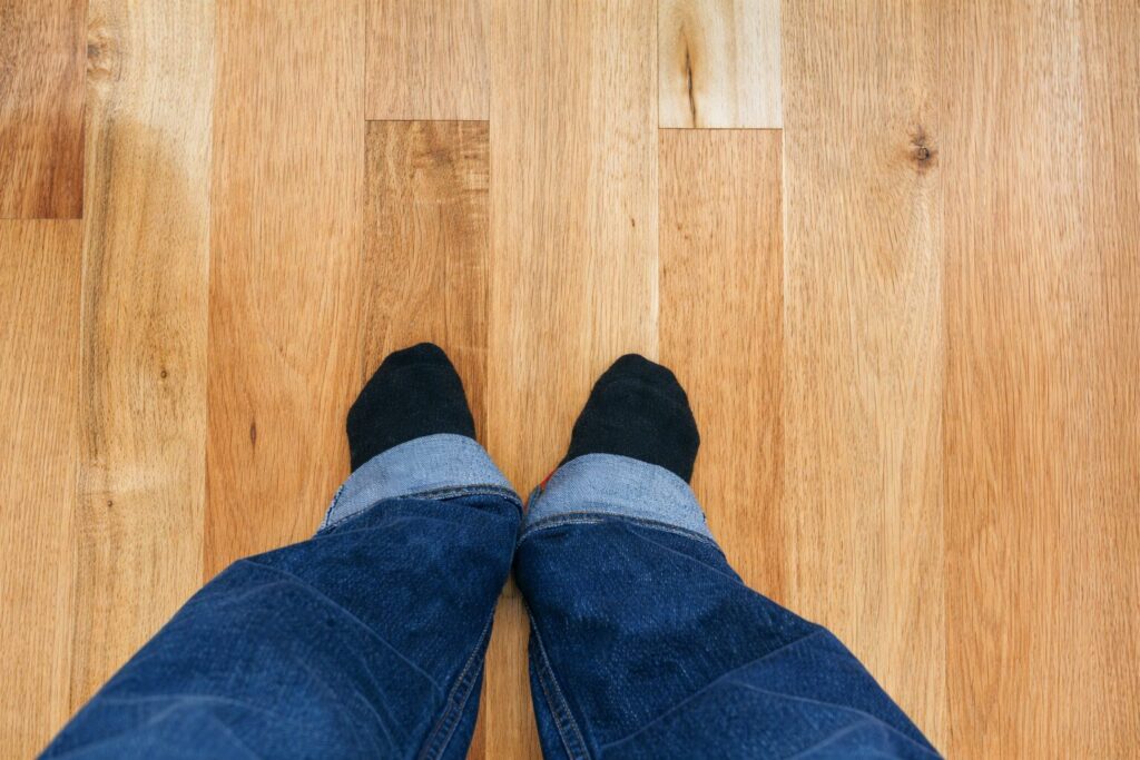 木目のあるフローリング床の上に、黒い靴下を履いた足先が並んで見える様子を上から撮影した構図。ジーンズの裾が写り込み、立った状態で床を見下ろしている視点が伝わる。