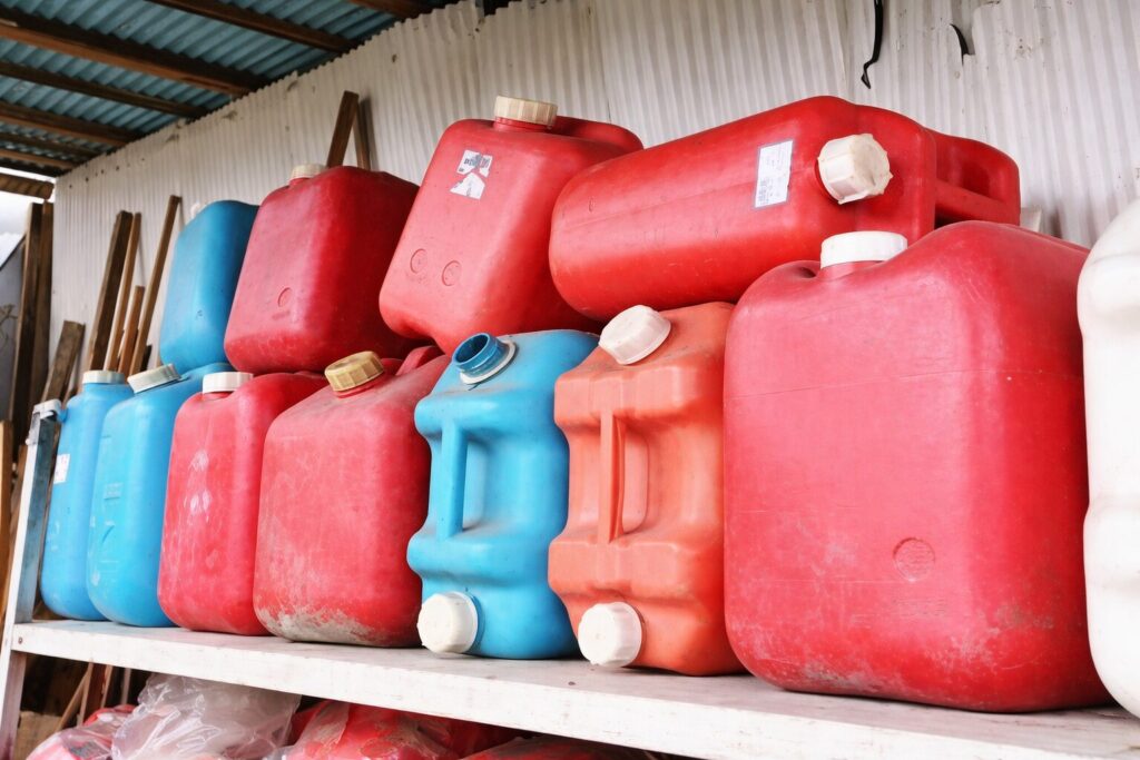 倉庫内の白い棚に、赤色と青色の灯油用ポリタンクが複数段に積み重ねて保管されている様子。屋根は波板で、使用感のあるポリタンクが整然と並んでいる。