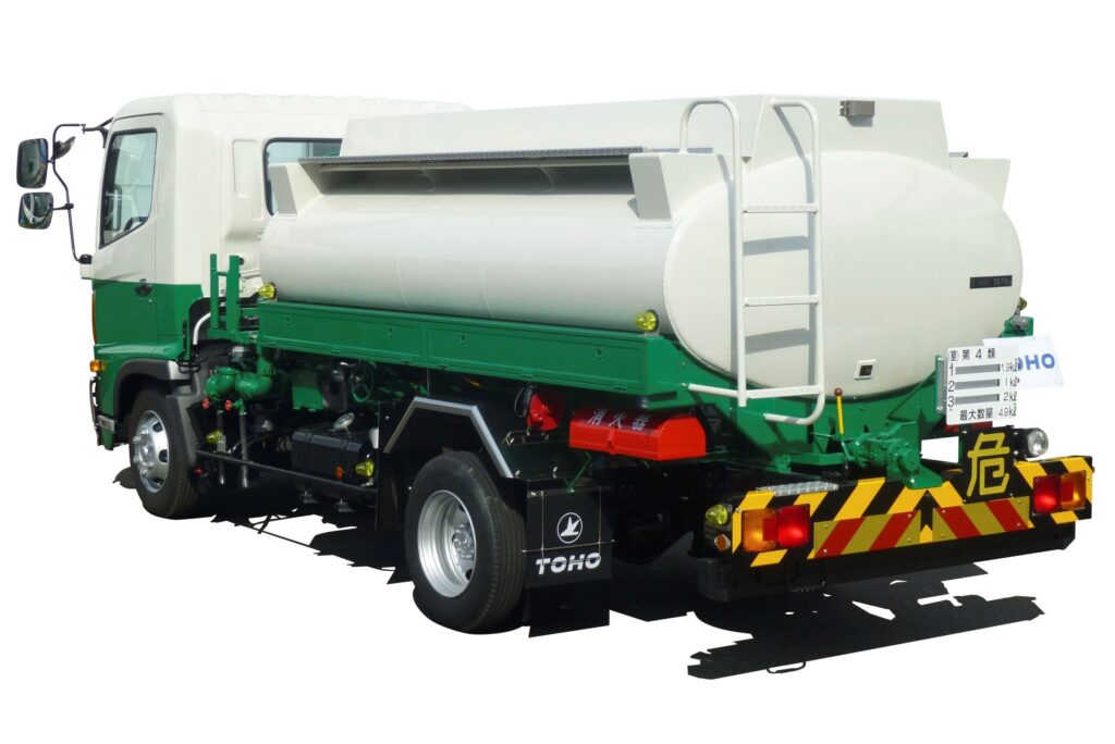 白色の円筒形タンクを荷台に搭載した灯油配送用のタンクローリー車両を、斜め後方から撮影した様子。緑と白のツートンカラーの車体で、後部には危険物表示と反射板が取り付けられており、家庭向け灯油配達に使用される専用車両の構造がわかる構図。