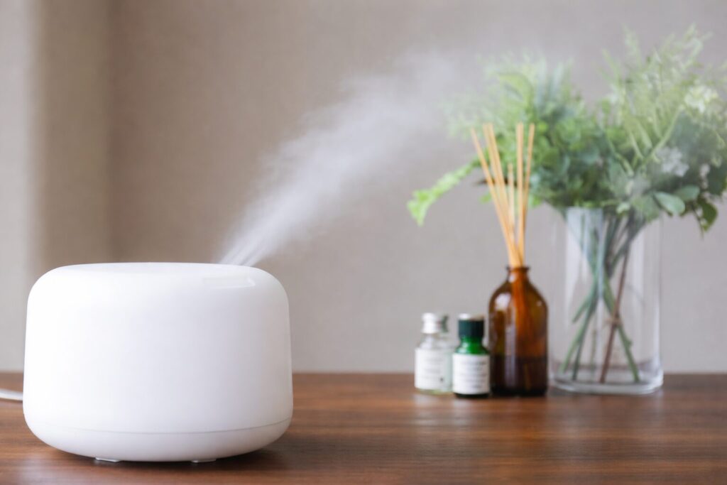 白い超音波式加湿器が木製テーブルの上に置かれ、上部の吹き出し口から細かな水蒸気が静かに広がっている様子。背後にはガラス花瓶のグリーンとアロマボトルが並び、室内の落ち着いた雰囲気が伝わる。