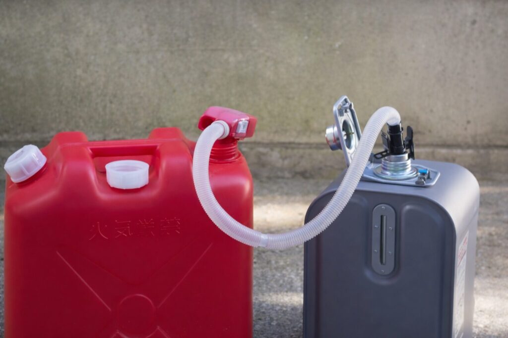 白い給油ホースで接続された赤い灯油用ポリタンクと灰色の金属製灯油タンクが、屋外のコンクリート上に並んで置かれ、灯油を移し替えている様子。