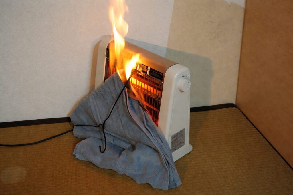 電気ストーブの前に掛けられたタオルが加熱され、炎が立ち上っている危険な状態を示す室内の様子。畳の床と壁際に設置されたストーブが写っており、可燃物との距離が近いことによる火災リスクが視覚的に伝わる構図。