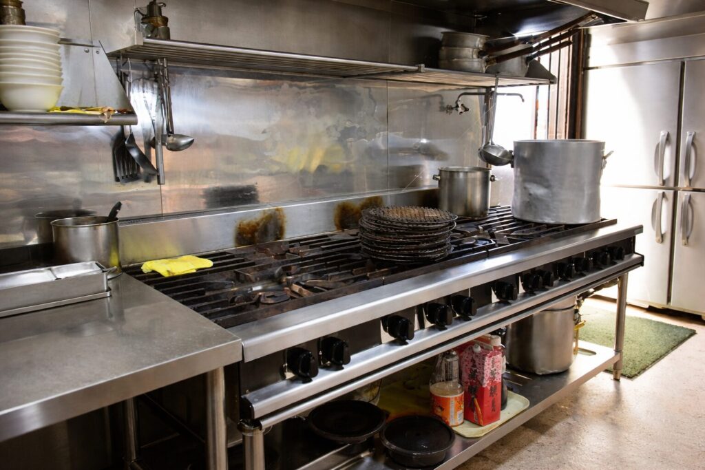業務用厨房に設置された大型ガスコンロを正面斜めから撮影した様子。ステンレス製の多口バーナーの上に寸胴鍋や小鍋、焼き網が並び、背面には油はねが付着したバックガードと、上部にはおたまやフライ返しなどの調理器具が吊り下げられている。下段の棚には調味料や鍋が収納され、実際に稼働している飲食店の厨房環境がわかる構図。