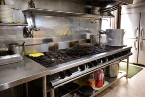 業務用厨房に設置された大型ガスコンロを正面斜めから撮影した様子。ステンレス製の多口バーナーの上に寸胴鍋や小鍋、焼き網が並び、背面には油はねが付着したバックガードと、上部にはおたまやフライ返しなどの調理器具が吊り下げられている。下段の棚には調味料や鍋が収納され、実際に稼働している飲食店の厨房環境がわかる構図。