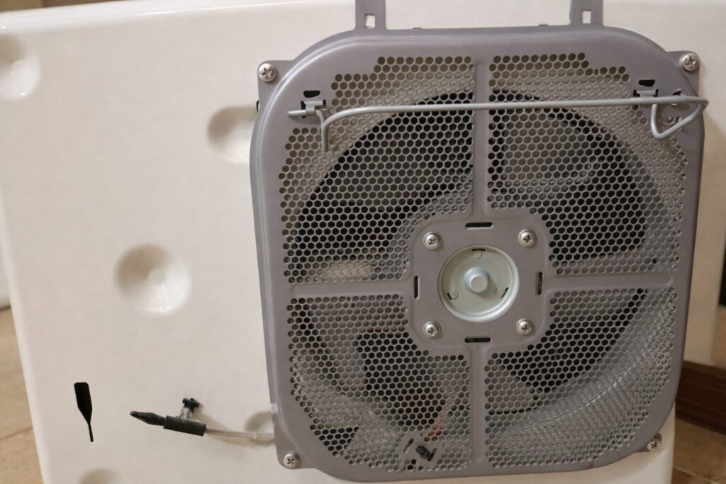 金属製のメッシュカバー越しに内部の羽根が見える換気ファンを、白い本体に取り付けた状態を背面から写した様子。配線や固定ネジも確認でき、内部構造が分かる構図。