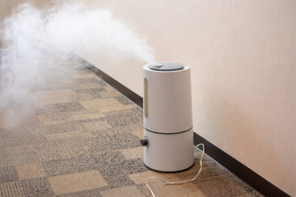 床に敷かれたカーペットの上に設置された円筒形の白い加湿器から、壁際に向かって白い蒸気が勢いよく噴き出している様子。