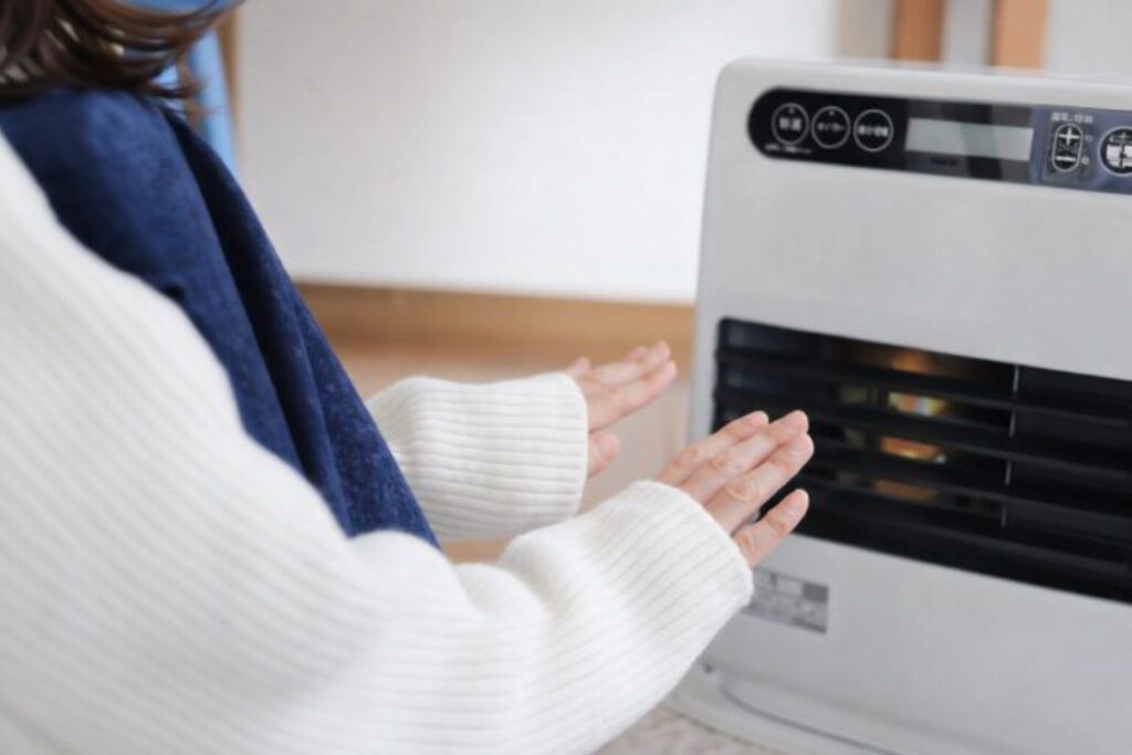 白いセーターを着た人物が、室内で白色のファンヒーターに両手をかざして暖を取っている様子。操作パネルが見えるヒーター前面から温風が出ており、落ち着いた生活空間の一場面を表している。