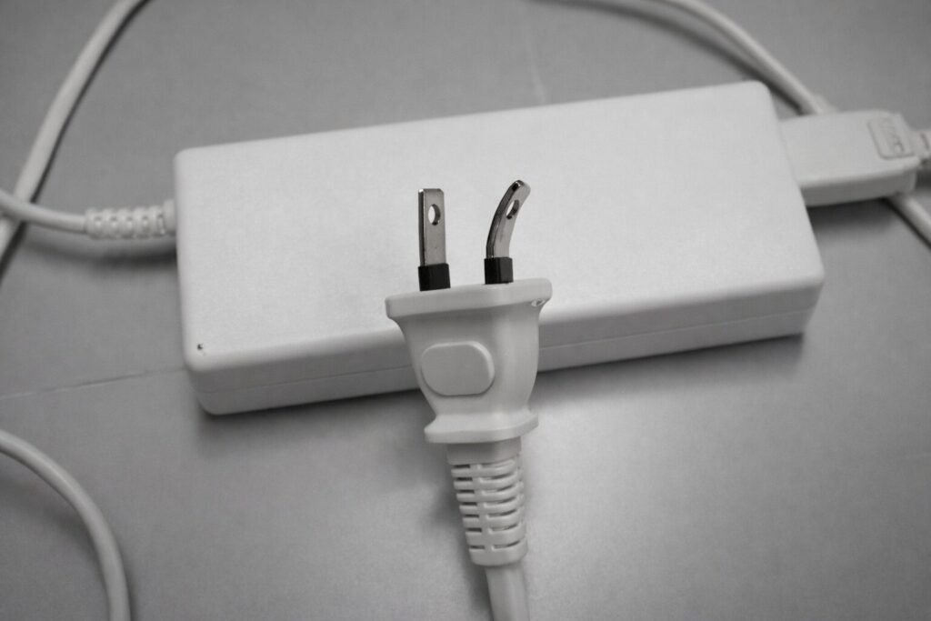 白い電源アダプターの前に、金属ピンの一方が曲がった電源プラグが置かれている様子。平らなグレーの面に横向きで配置され、プラグの変形状態が分かる構図。