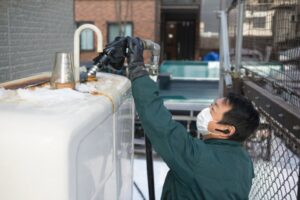 雪が積もった屋外の灯油タンク上部で、作業着と手袋、マスクを着用した配達員が給油ホースを接続し、灯油を補給している様子。住宅設備が並ぶ建物脇の通路で行われている冬季の灯油補給作業がわかる構図。