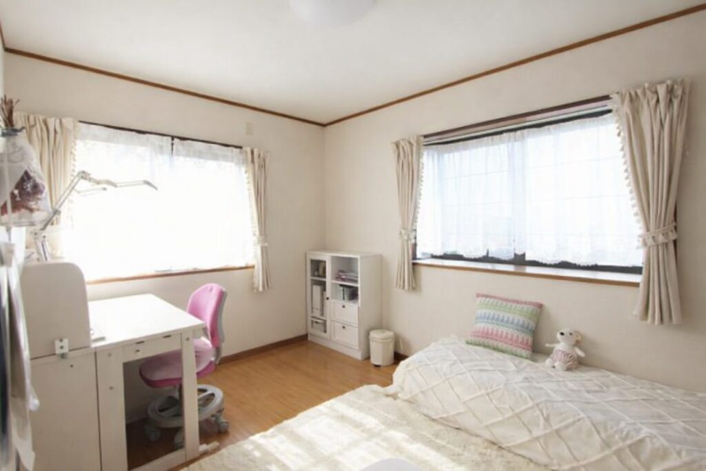 自然光が入る明るい個室の様子。白を基調とした部屋に、窓際に配置された白い学習机とピンク色の椅子、シンプルなベッドとクッション、小型の収納棚が並び、整理された落ち着いた室内空間が広がっている。