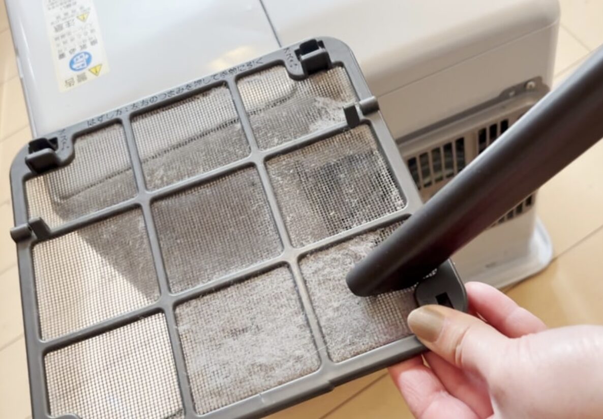 ファンヒーターの吸気フィルターを取り外し、掃除機のノズルで網目に詰まったホコリや綿ぼこりを吸い取っている様子。背景には本体の吸気口が見える。