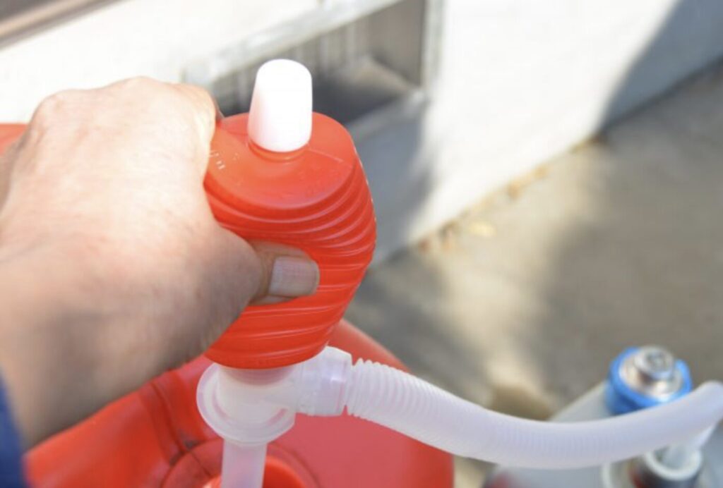 灯油タンクに取り付けた赤色の手動式灯油ポンプを手で押して給油している様子。白いホースが接続され、屋外で作業している場面が確認できる。