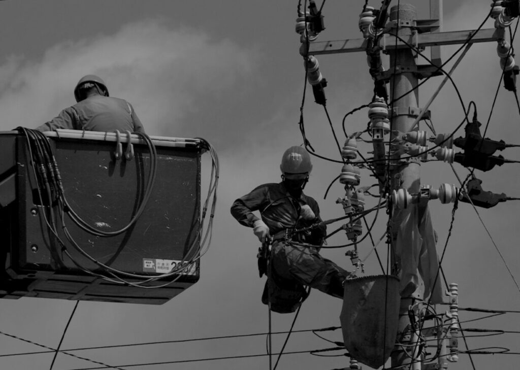 電柱に取り付けられた電力設備の点検作業を行う作業員が、高所作業車と安全装備を使用して作業している様子