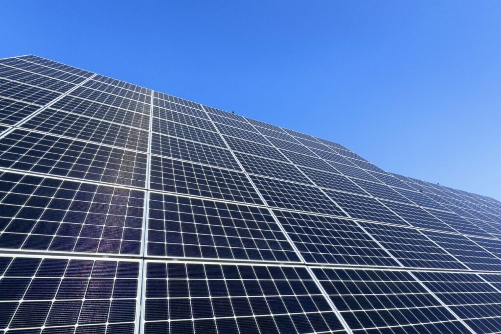 青空の下に広がる太陽光パネルの表面を、斜め下から見上げる構図で捉えた発電設備の様子
