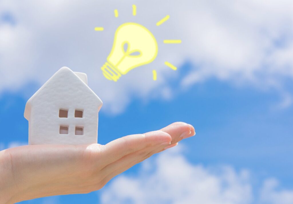 手のひらに小さな白い家の模型を載せ、その上に電球のアイコンが浮かび、住まいと電気エネルギーの関係性を表現したイメージ
