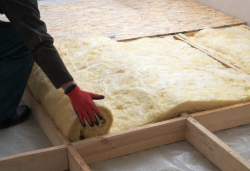 床下地の木枠の間に、ロール状のグラスウール断熱材を手作業で敷き込み、隙間が出ないよう調整しながら施工している様子を写した写真。