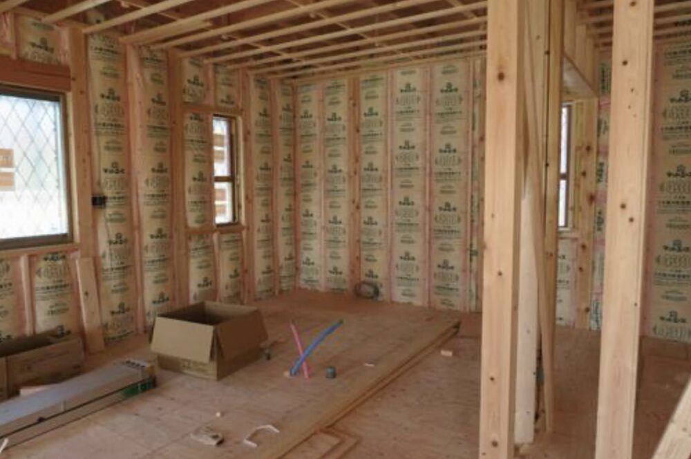 木造住宅の新築工事中の室内の様子。柱や梁が露出した状態で、壁と天井には断熱材が隙間なく施工され、床には配管材や段ボール箱などの建築資材が置かれている内装前の空間。