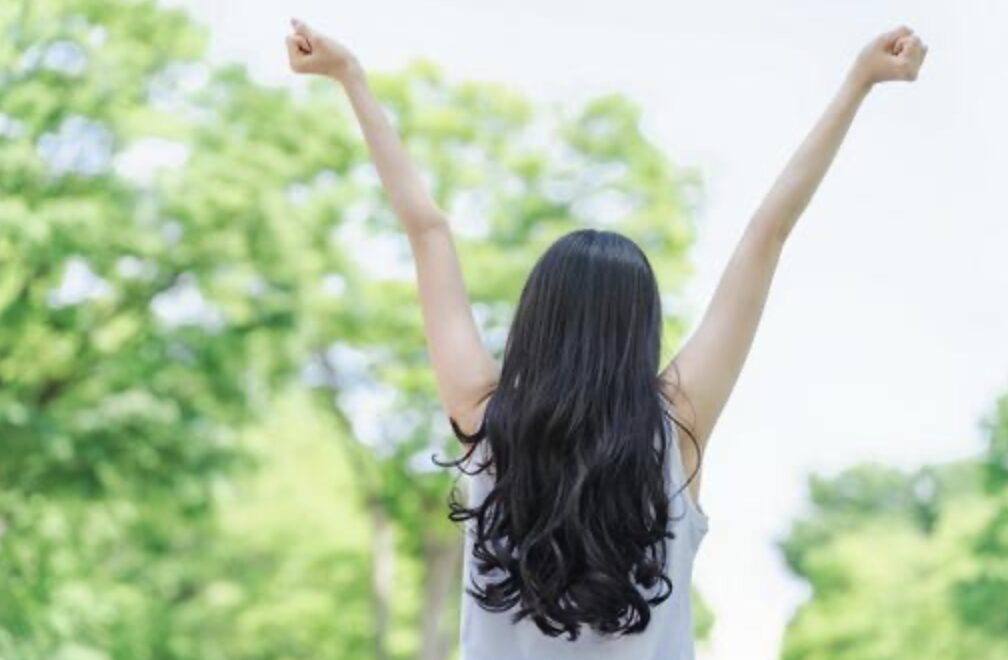 緑の木々が広がる屋外で、長い黒髪の人物が両腕を大きく上に伸ばし、背中を向けて伸びやかに体を動かしている様子