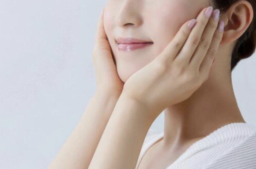 清潔感のある明るい背景の中で、人物が両手で頬に触れ、肌のうるおいやなめらかさを確かめている様子