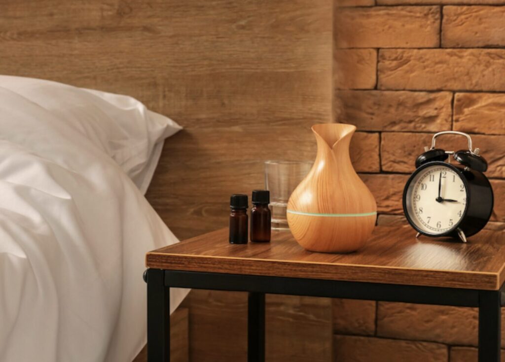 寝室のベッド脇にある木製サイドテーブルの上に、木目調の加湿器と目覚まし時計、アロマボトルが並び、落ち着いた雰囲気を演出している様子