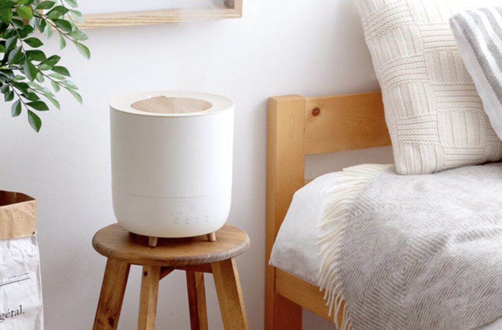 ベッド脇の木製スツールの上に設置されたシンプルなデザインの加湿器が、明るく清潔感のある寝室になじんで配置されている様子