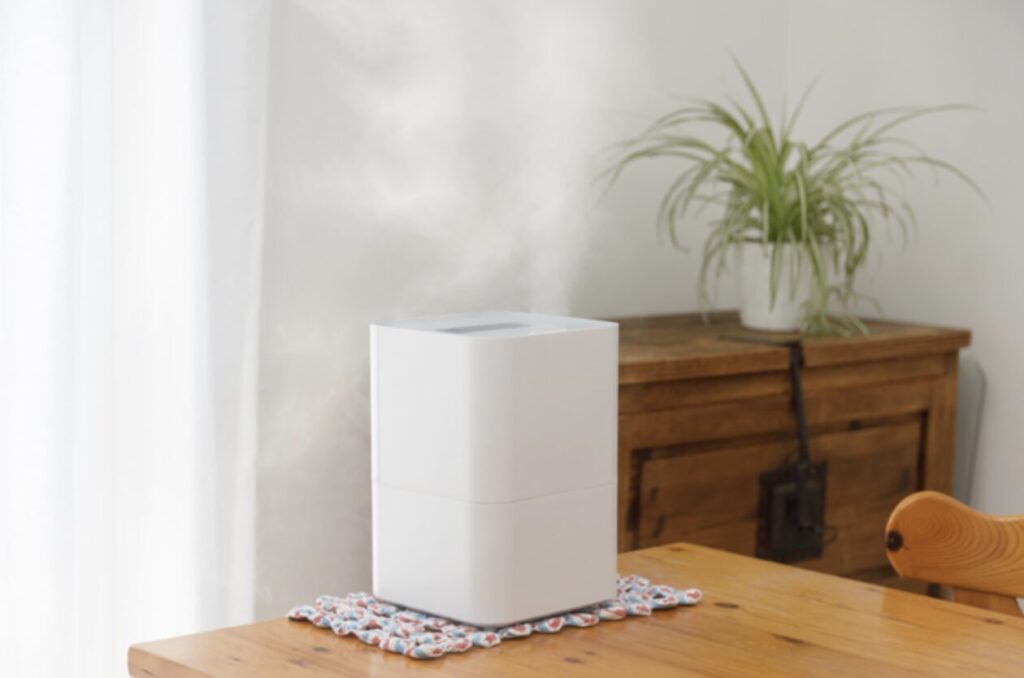 木製テーブルの上に設置された卓上型の加湿器から、やわらかな白いミストが立ち上り、背後には観葉植物と収納家具が配置された落ち着いた室内空間の様子