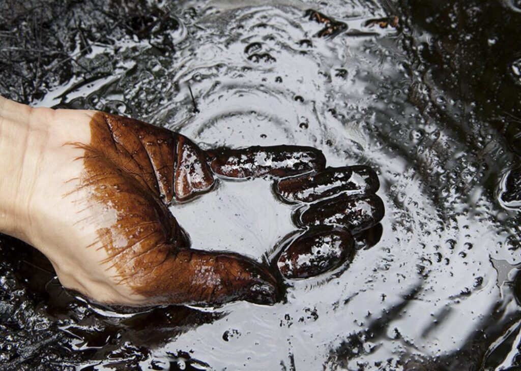 石油を手ですくい上げている様子で、手のひらや指に粘り気のある油分が付着している状態。石油や油汚れが土や水と混ざった状況がわかる。