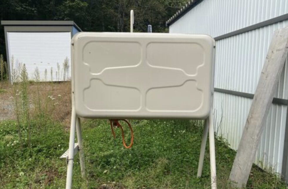 屋外に設置された家庭用の灯油タンクを正面から撮影した様子で、脚付きのタンク本体と下部に接続された配管が確認できる。物置や外壁の近くに設置されており、住宅での灯油保管環境がわかる。