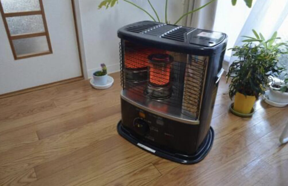 木目のフローリングの室内に設置された据置型の石油ストーブが燃焼している様子で、前面ガード越しに赤く発光する燃焼筒が確認できる。周囲には観葉植物が置かれ、住宅のリビング空間で使用されている状況がわかる。