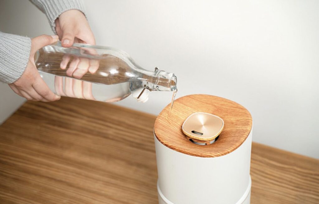 手に持ったガラスボトルから、水を上から注いで加湿器本体に給水している様子。木目調の天板とシンプルな円筒形デザインが確認できる。