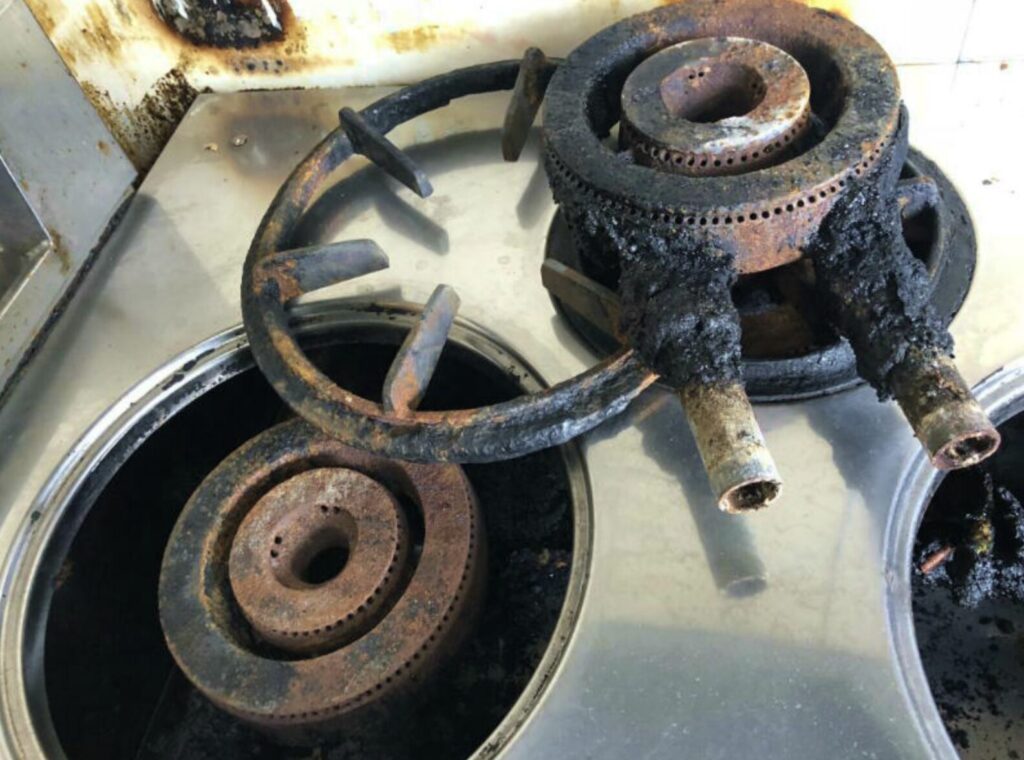 分解された業務用ガスコンロのバーナーと五徳を並べた状態。バーナー本体やガス噴出口周辺に炭化した油汚れやサビが目立ち、清掃前の内部構造がわかる構図。