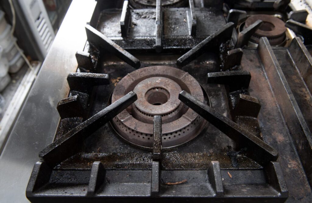 業務用ガスコンロの五徳とバーナーを正面から捉えた様子。鋳物製五徳の隙間やバーナー周囲に黒い油汚れや焦げが蓄積しており、使用頻度の高い厨房設備の状態が伝わる。