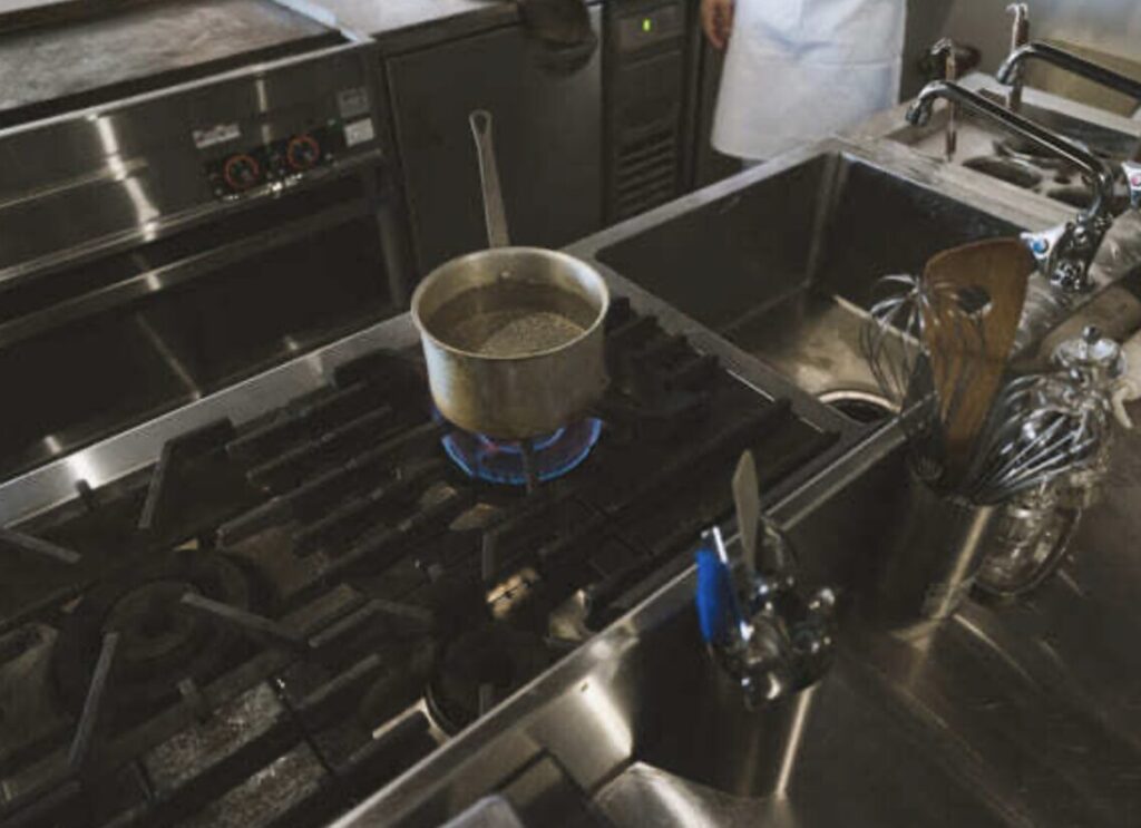 業務用ガスコンロの上で鍋を加熱している様子。青いガスの炎がバーナーから立ち上がり、隣には業務用シンクや調理器具が配置されている厨房内の実使用シーン。