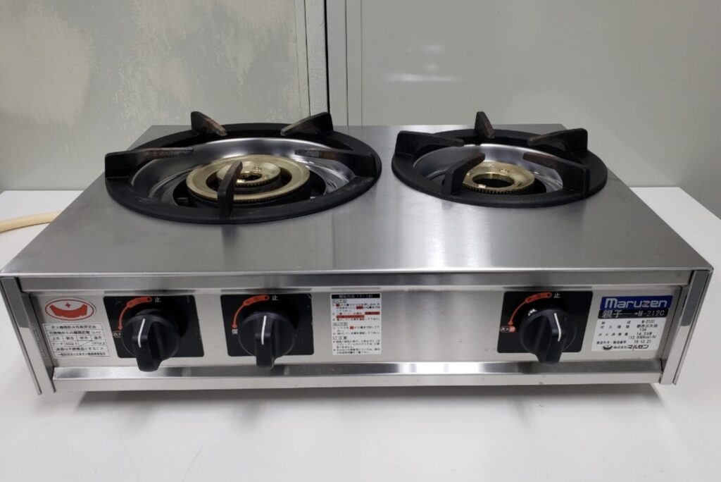 ステンレス製の卓上型業務用ガスコンロを正面から撮影した様子。2口の鋳物製バーナーと前面に配置された操作ノブが確認できるシンプルな構造。