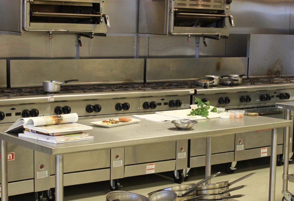 業務用厨房に設置された大型のガスコンロとオーブンレンジが横一列に並ぶ様子。ステンレス製の調理台の上には鍋や食材、調理器具が配置され、プロの調理環境が伝わる構図。