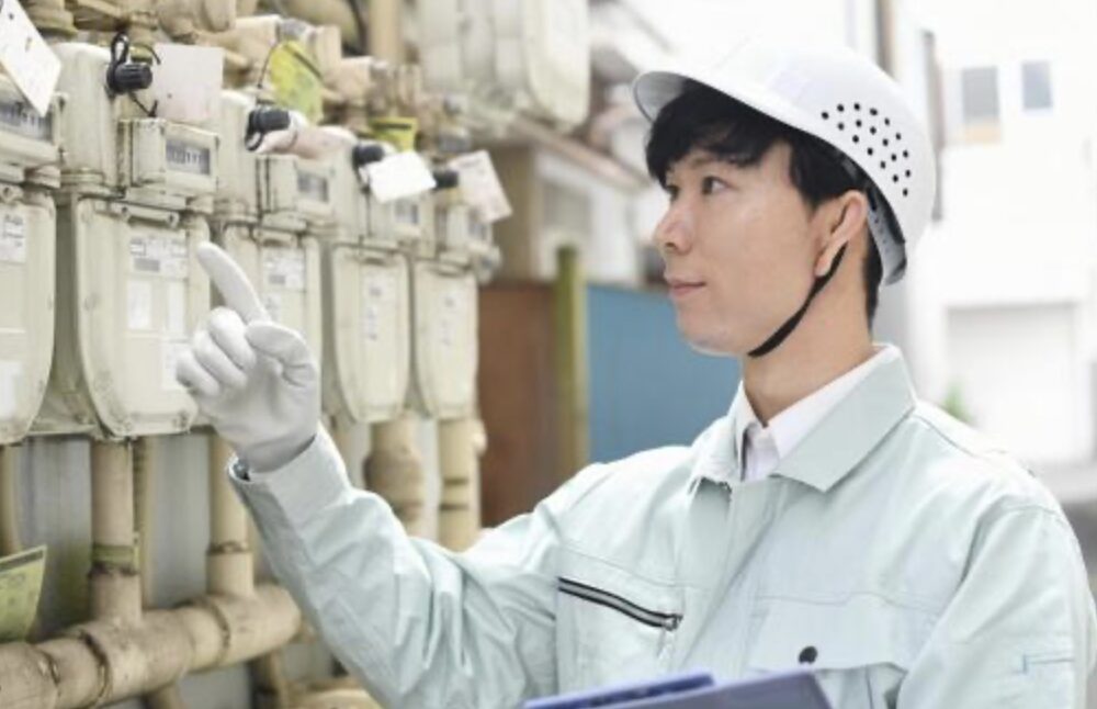 屋外に設置された複数のガスメーターを前に、保安帽と作業着を着用した点検員が指差し確認を行っている様子。検針や保安点検の作業風景がわかる構図。