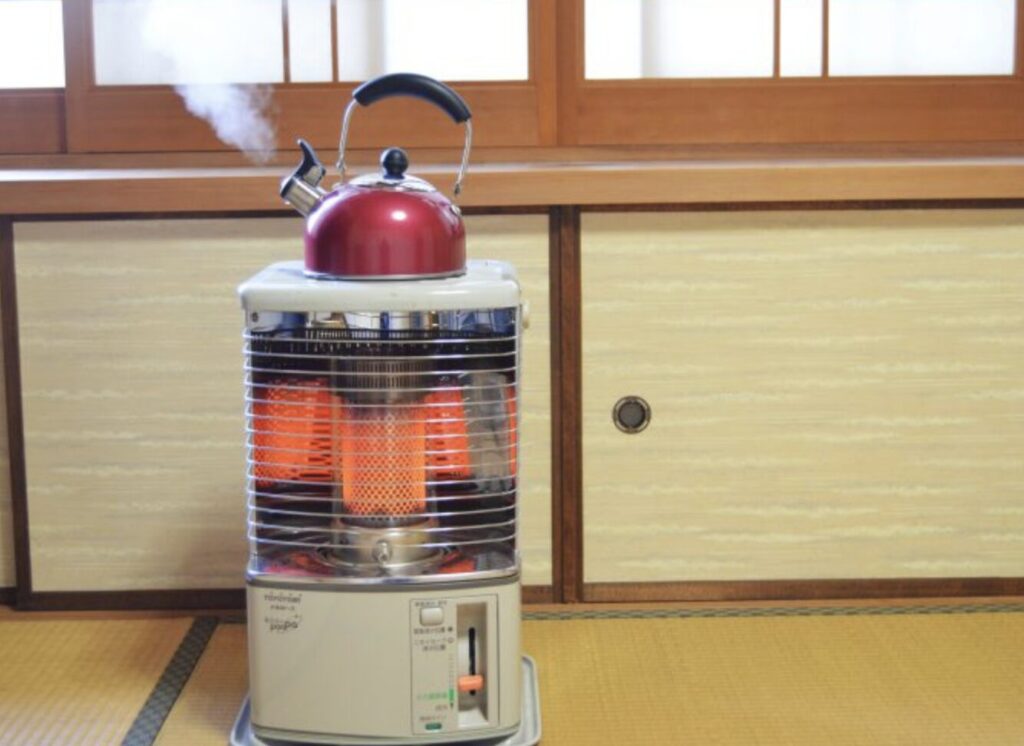 和室の畳の上に設置された反射式の石油ストーブの上で、赤いやかんから湯気が立ち上っている様子。ストーブ内部の燃焼筒が赤く発光し、障子と木製建具に囲まれた室内で暖房と湯沸かしを兼ねて使用されている状況がわかる構図。