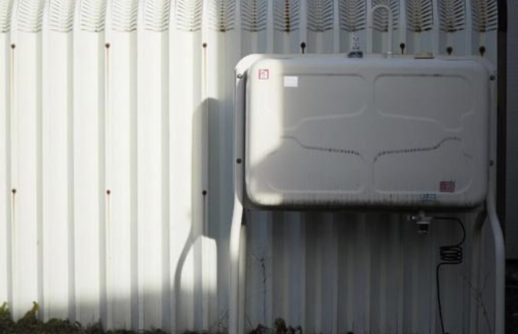 外壁の波板金属パネルに沿って設置された屋外用の灯油タンクを、正面から横長構図で撮影した様子。白色の角型タンク本体と配管が確認でき、日差しによってタンクと配管の影が壁面に落ちている屋外設置状況がわかる構図。