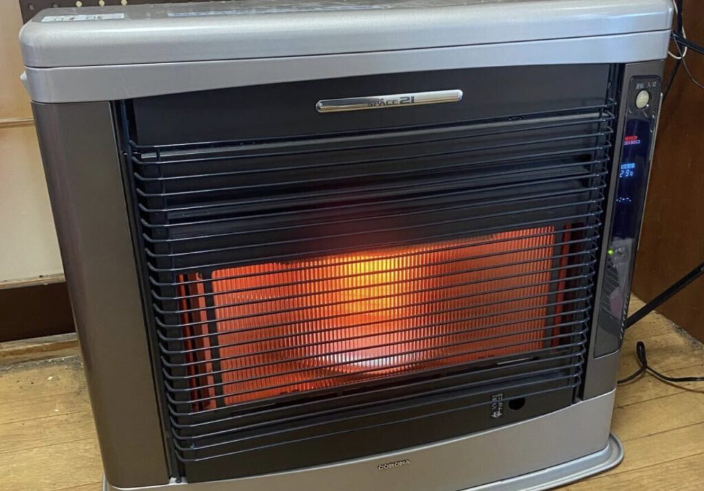 室内に設置されたコロナ製FF式石油ストーブの正面外観。前面ガード越しに燃焼中のオレンジ色の炎が見え、本体上部に操作パネルが配置されている。