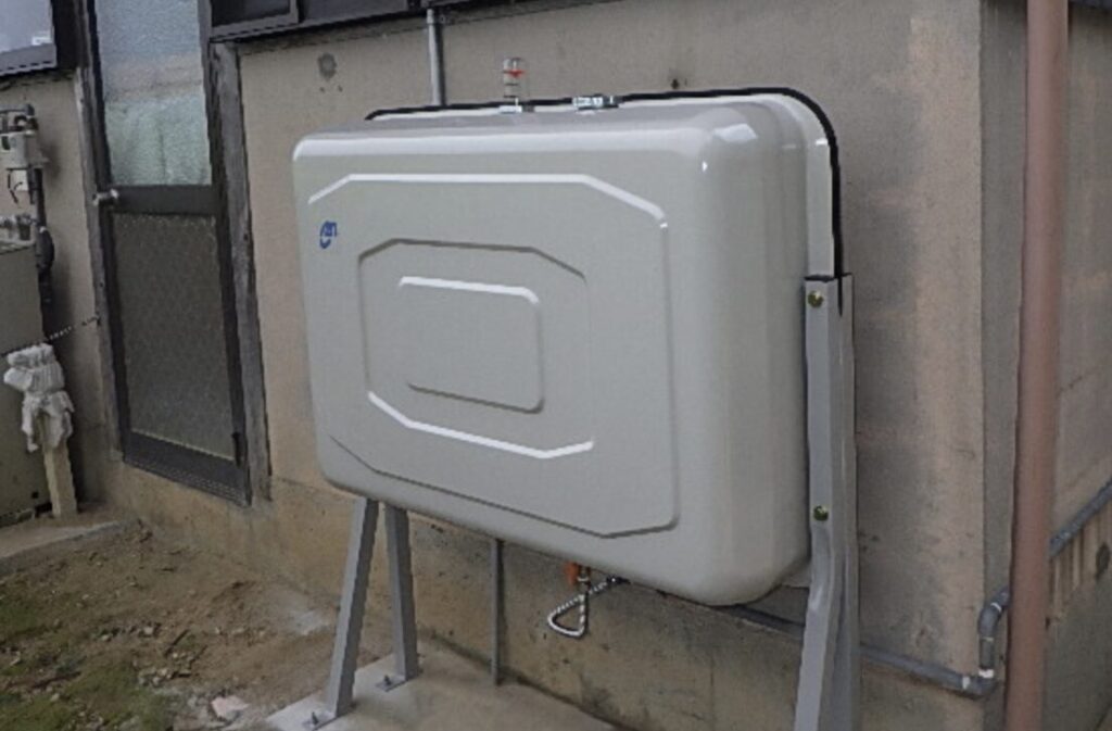 住宅外壁のそばに設置された灯油タンクを斜めから撮影した様子。配管接続部と支持金具の位置関係が確認でき、屋外設置の状態がわかる。