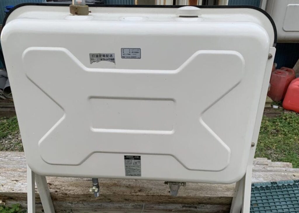 屋外に設置された角型の灯油タンクを正面から撮影した様子。白色の金属製タンクが専用架台に固定され、上部に給油口と通気口が配置されている。