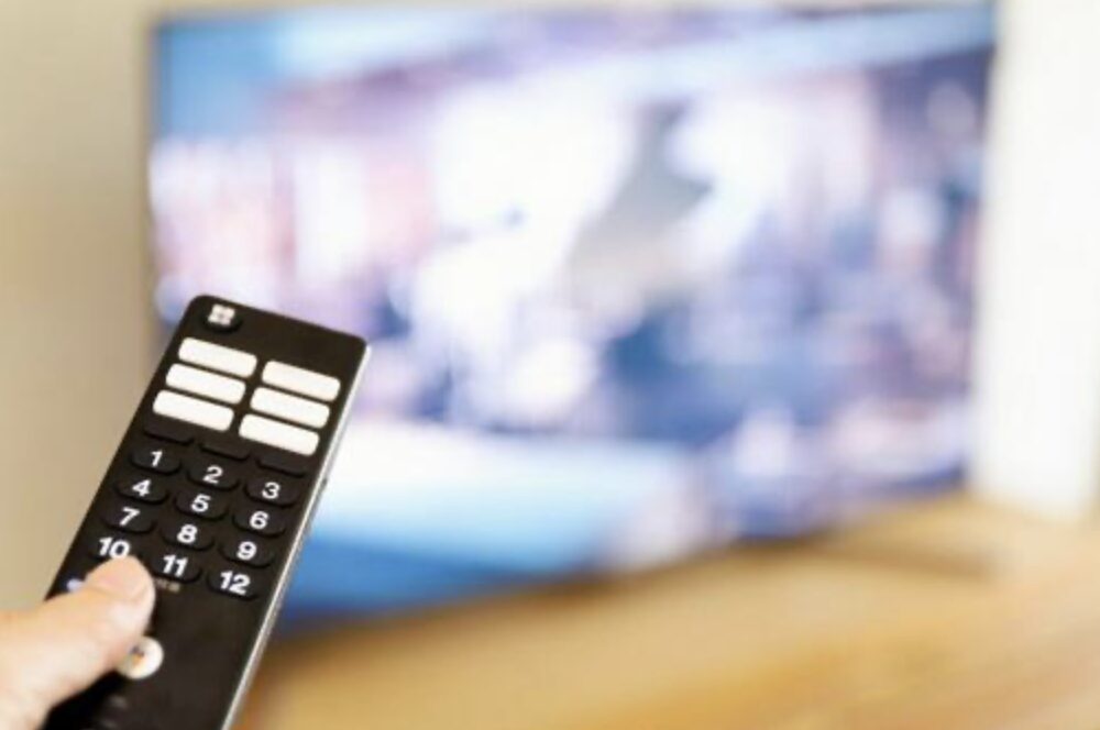 テレビ画面に向かって手に持ったリモコンを操作している様子で、室内での視聴シーンがぼかして写っている。