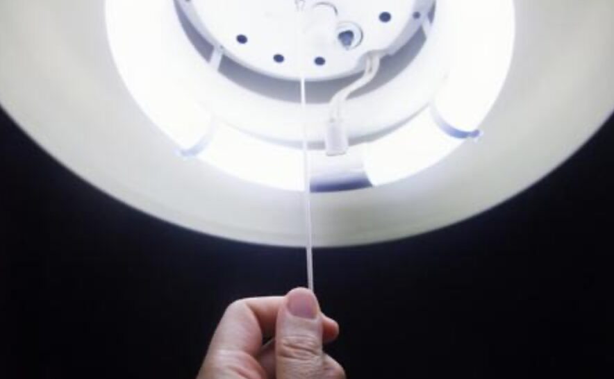 天井照明の紐スイッチを手で引いて点灯・消灯操作をしている様子。照明内部の蛍光灯が点灯している。